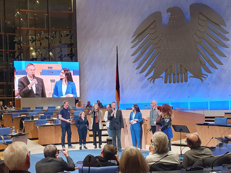 Blick ins Plenum, wo Politiker_innen im Gespräch stehen mit Mikrofon, im Hintergrund werden sie auf Leinwand übertragen, im Vordergrund des Bildes sitzen die Zuschauer