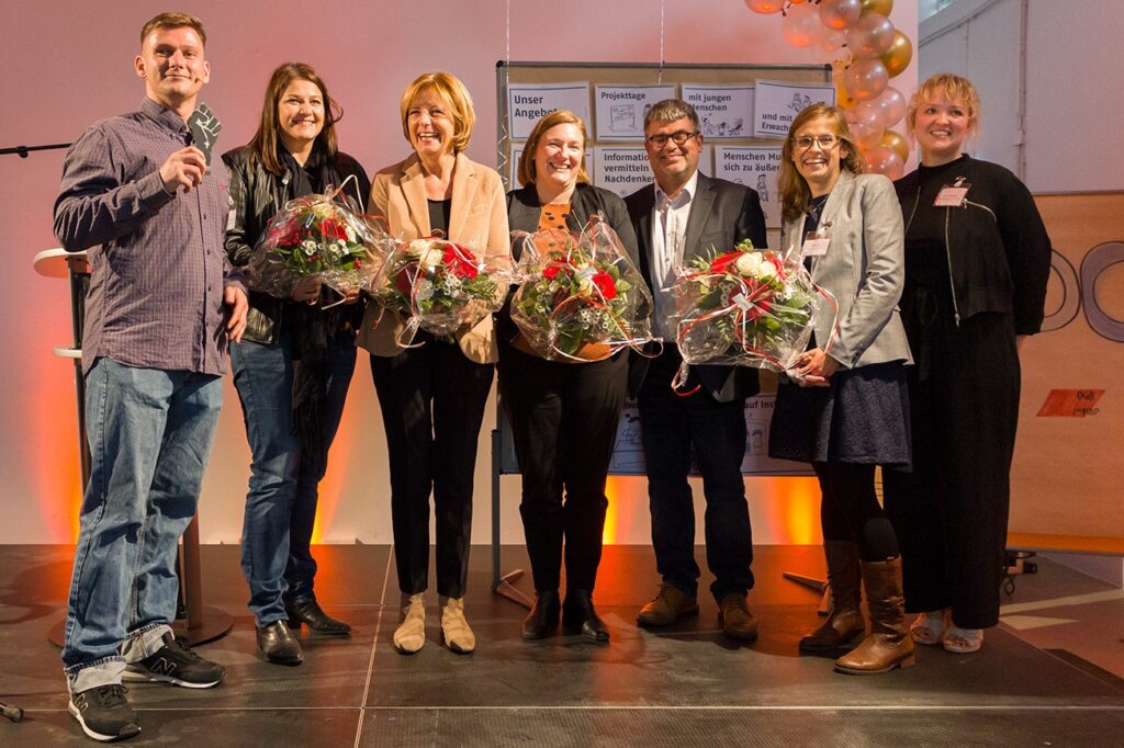 7 Menschen stehen nebeneinander, z.T. halten sie Blumensträuße in der Hand.