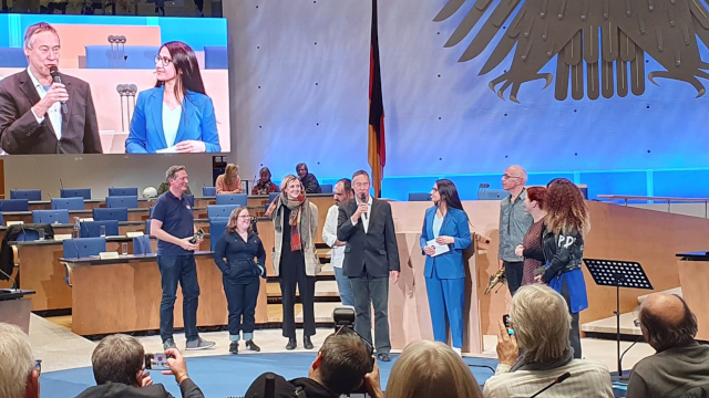 Blick ins Plenum, wo Politiker_innen im Gespräch stehen mit Mikrofon, im Hintergrund werden sie auf Leinwand übertragen, im Vordergrund des Bildes sitzen die Zuschauer