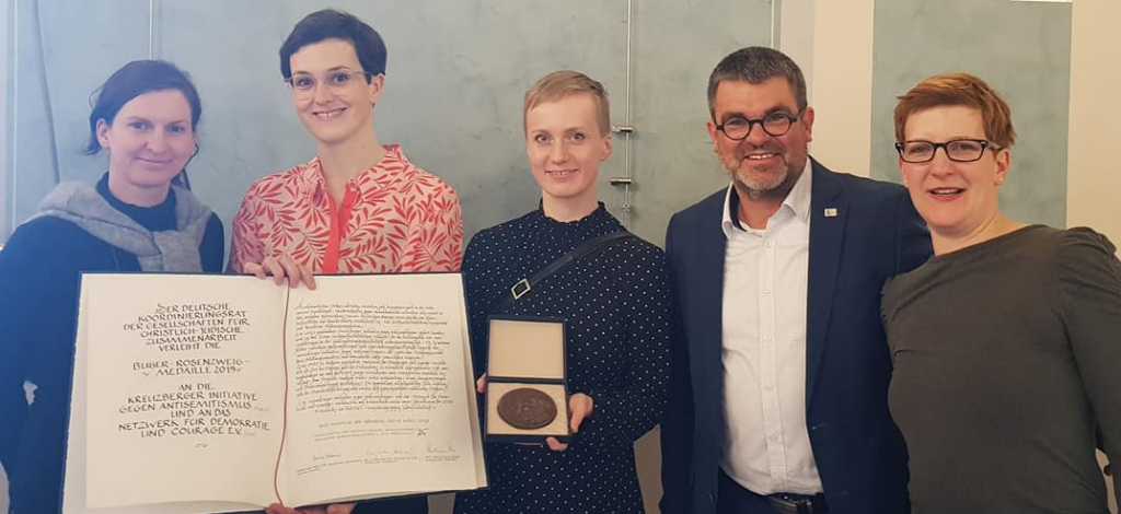 Auf dem Bild sind 5 Personen zu sehen, zwei davon halten die Urkunde der Buber-Rosenberg-Auszeichnung für das NDC in der Hand, eine Person hält die Medaillie in der Hand. Alle freuen sich.
