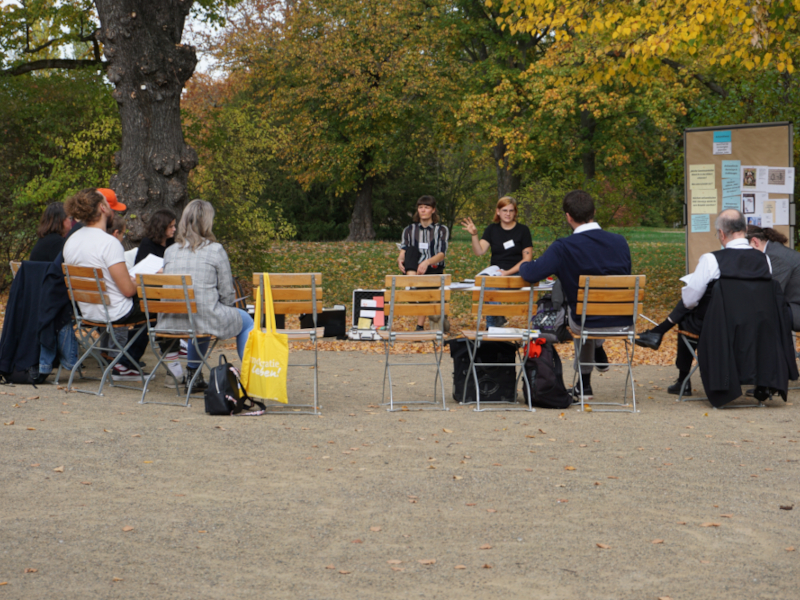 Mehrere Personen sitzen in einem Stuhlkreis, der im Freien unter einem Baum aufgebaut ist. Neben den zwei Workshop-Leiterinnen steht eine Pinnwand und ein offener Moderationskoffer.