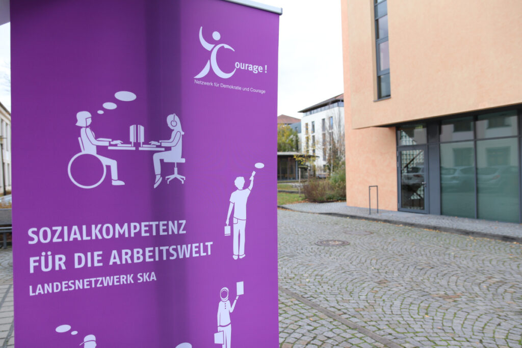 Im Vordergrund ein Roll-up mit der Aufschrift "Sozialkompetenz für die Arbeitswelt - Landesnetzwerk SKA", im Hintergrund ein Gebäude