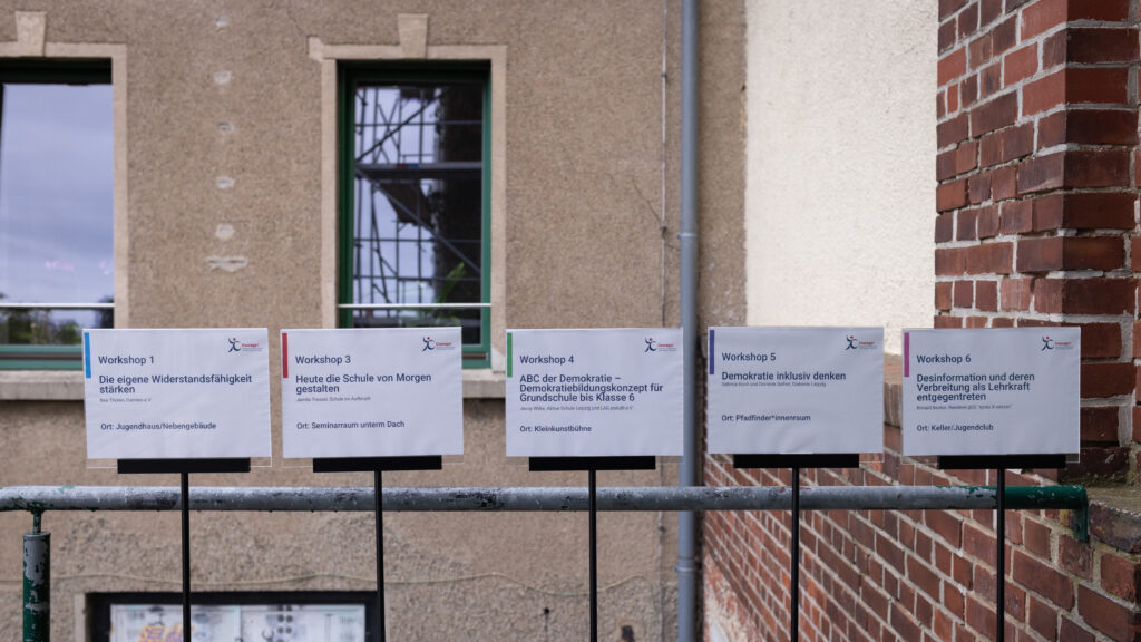 An einem Geländer in einem Innenhof sind fünf Schilder befestigt, auf denen Workshopbeschreibungen stehen