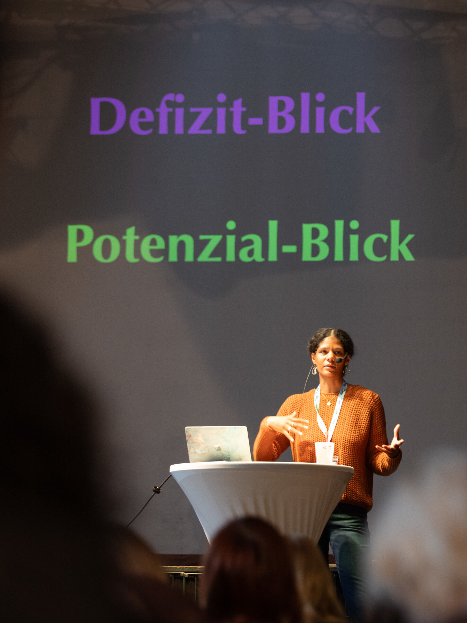 Jamine Dressel spircht als Referentin und steht am Rednerpult. Vor ihr ein aufgeklapter Laptop. Hinter ihr stehen die Worte Defizit-Blick und Potenzial-Blick