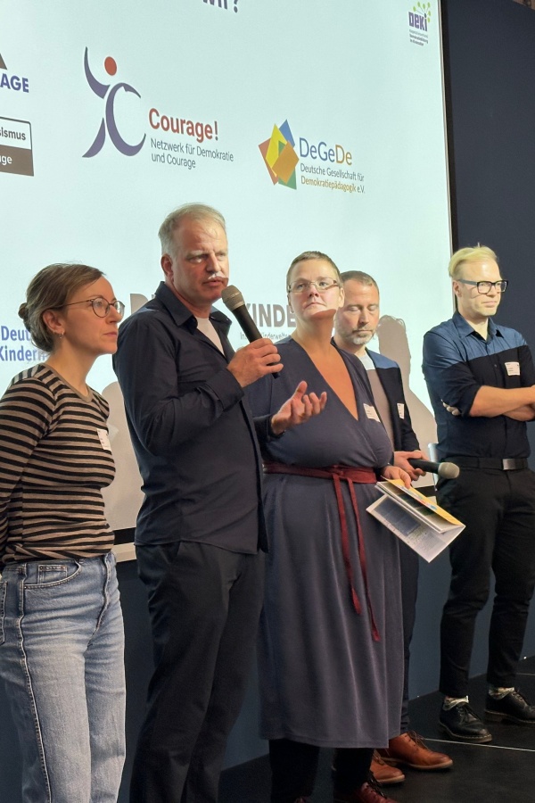 Fünf Vertreter der Trägerorganisationen stehen auf der Bühne. Andreas Stäbe spricht ins Mikrofon. Im HIntergrund sind die Logos des NDC und der DeGeDe zu sehen.