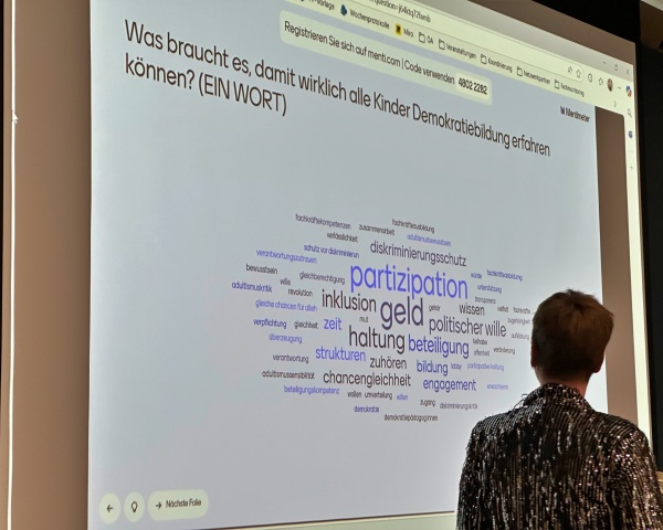 Eine Person blickt auf eine an die Wand projizierte Wortwolke. Über der Wortwolke steht die Frage: Was braucht es, damit wirklich alle Kinder Demokratiebildung erfahren können? Die prominentesten Begriffe sind: Partizipation, Geld, Haltung, Beteiligung, politischer Wille und Inklusion.