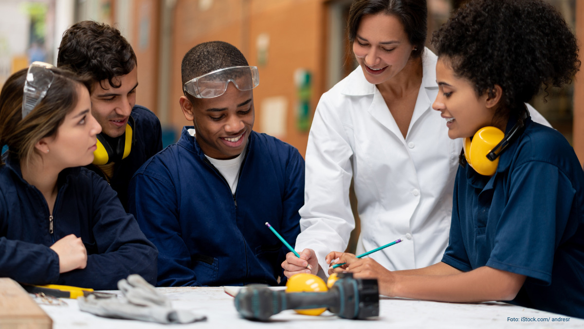 Menschen in Berufskleidung arbeiten an einer Zeichnung, es liegen auch ein Akkuschrauber, Handschuhe, Zange und Gehörschutz auf dem Tisch