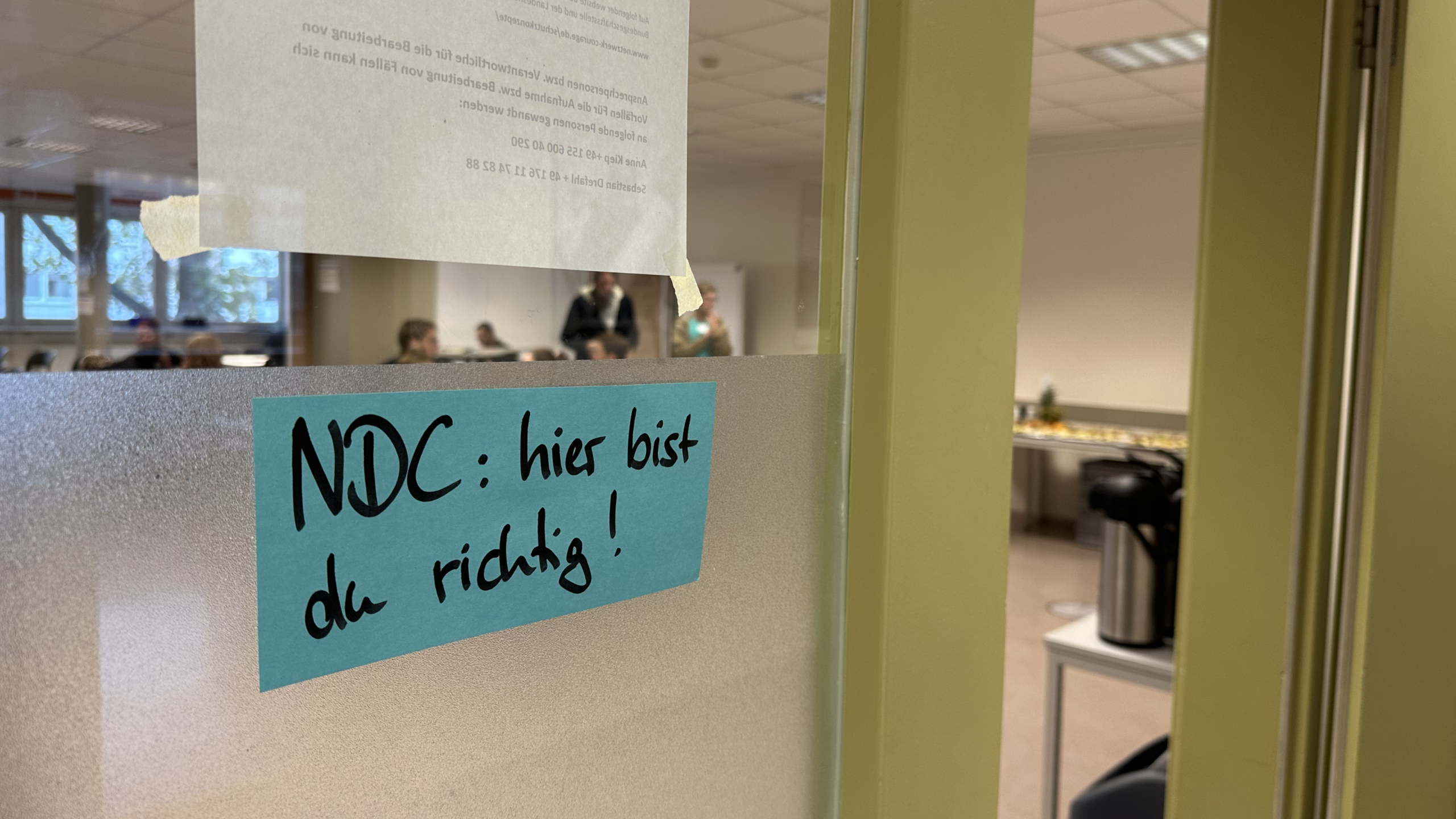 Eine Glastür, an der ein Zettel mit der Aufschrift: "NDC: hier bist du richtig! hängt". Im Hintergrund ein Konferenzraum mit einigen Menschen und Tischen mit Verpflegung und Kaffee.