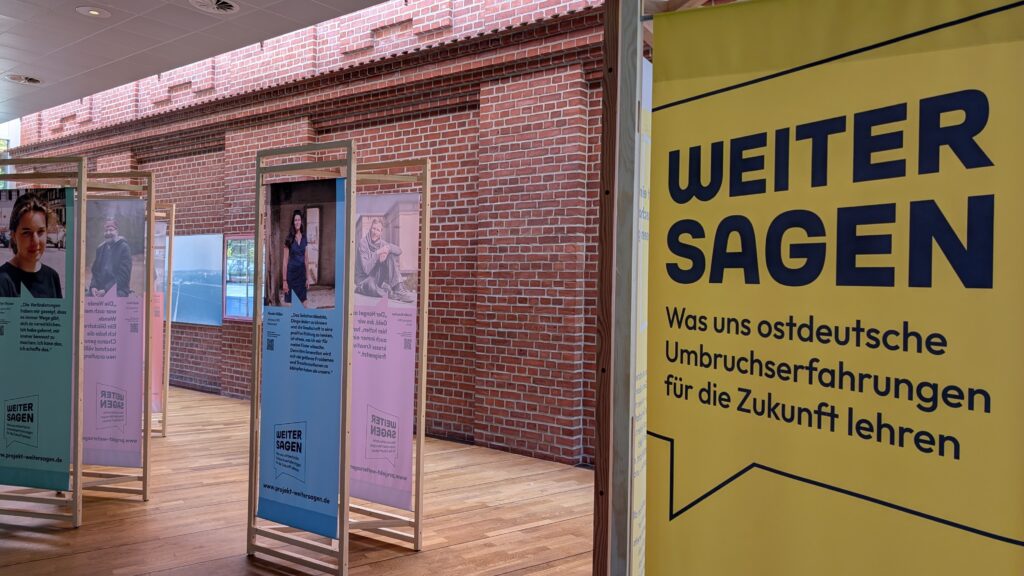 In einem großen Raum stehen mehrere hochformatige Stellwände, die mit Fotos und Texten die Ergebnisse des Projektes Weitersagen zeigen. Im Vordergrund kann man auf einer Stellwand lesen: Weitersagen: was uns ostdeutsche Umbruchserfahrungen für die Zukunft lehren.