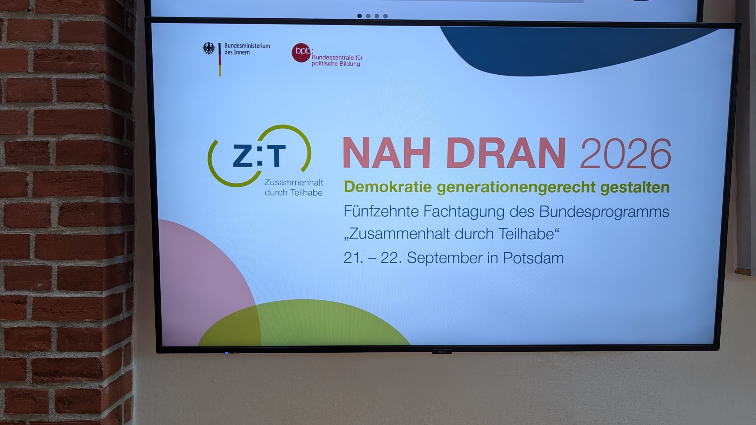Ein Flatscreen hängt an einer Wand. Auf dem Bildschirm sieht man das Logo des Bundesprogramms Zusammenhalt durch Teilhabe. Daneben steht: Nah Dran 2026. Demokratie generationsgerecht gestalten. 15. Fachtagung des Bundesprogramms "Zusammenhalt durch Teilhabe", 21.-22. September in Potsdam.